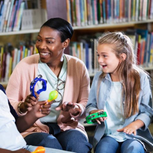 A group of three multiracial children and a women, mother of the two boys, sitting together in a school library making music with toy tambourines and maracas. The children are 8 to 11 years old.